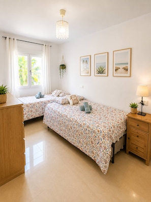 A bedroom with two beds, a hanging plant, framed pictures on the wall, and a lamp on a bedside table. - Spacious 2BR Pool Parking Near the Beach (Alicante)