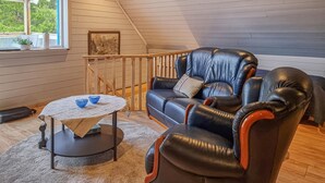 A cozy room with a wooden ceiling, a black leather sofa, a small round table with a white tablecloth, and a window with a view of greenery.