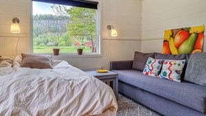 Ett sovrum med en säng, en grå soffa, ett fönster med utsikt över grönområden och två vägglampor.