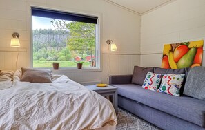 A bedroom with a bed, a gray sofa, a window with a view of greenery, and two wall-mounted lamps. - Nice home in Vänersnäs with house a mountain view (Vänersnäs)