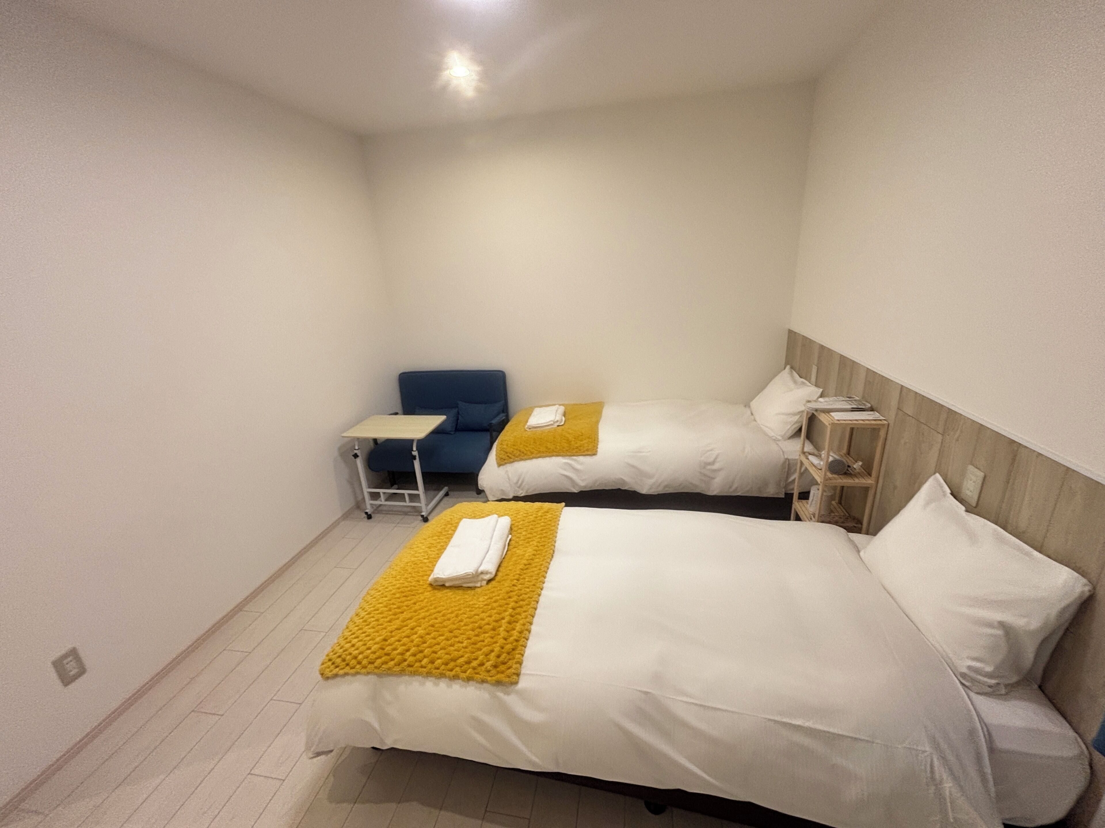 A hotel room with two beds, a small table, a blue chair, and a wooden shelf.