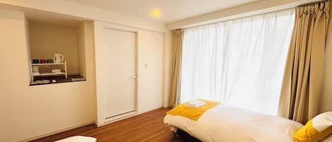 Quarto de hotel com uma cama, um pequeno espaço de estar, uma prateleira com objetos e uma janela com cortinas.