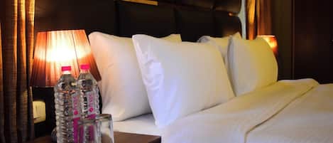 A hotel room with a bed, pillows, a nightstand with water bottles, and a lamp.