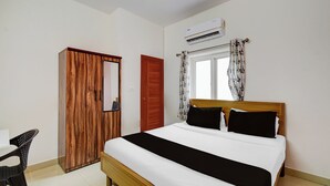 Ein Schlafzimmer mit einem Holzschrank, einem Bett mit schwarz-weißer Bettwäsche, einem Fenster mit blumigen Vorhängen und einer Klimaanlage.