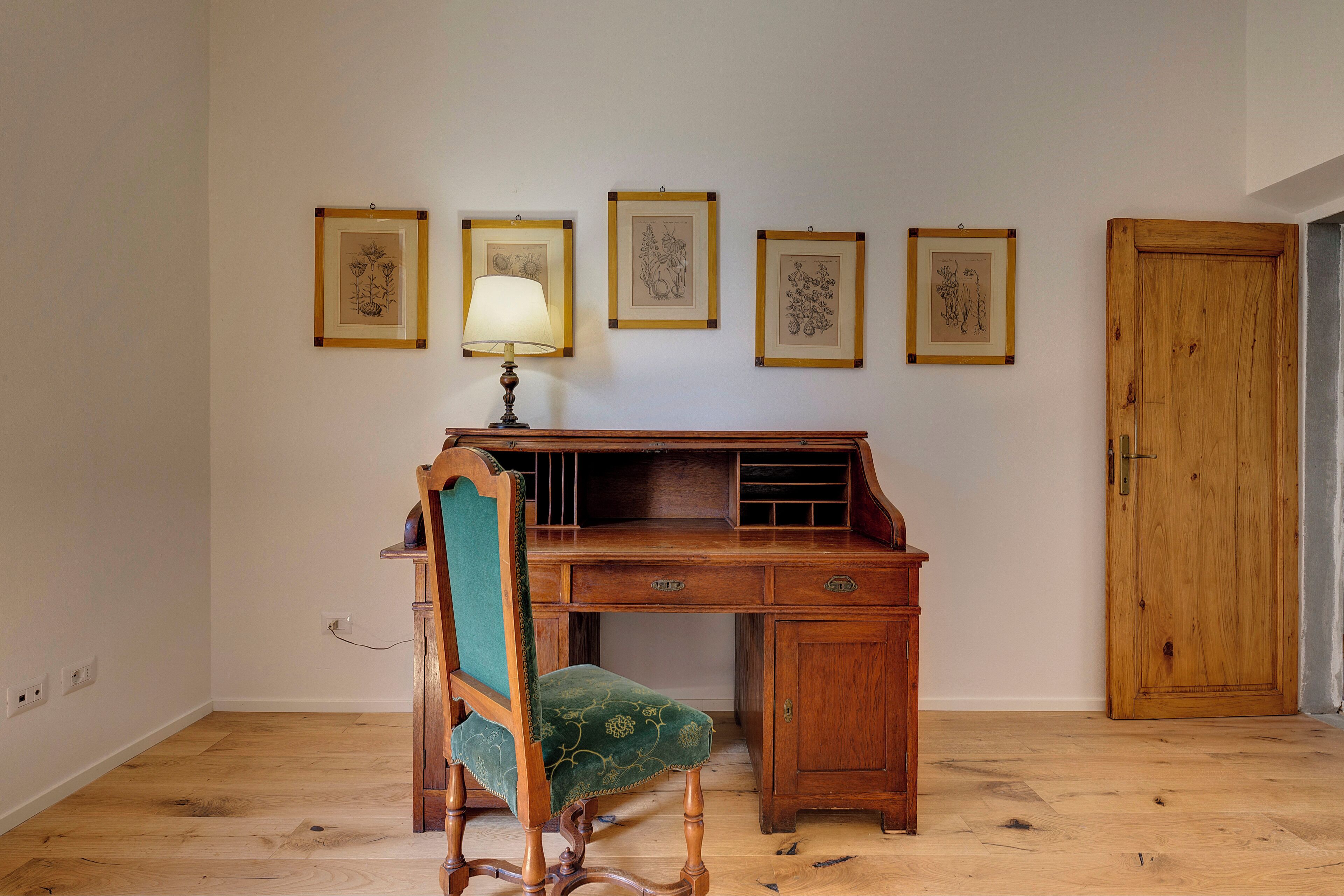 Un escritorio de madera con una silla, una lámpara y cuatro ilustraciones botánicas enmarcadas en la pared.