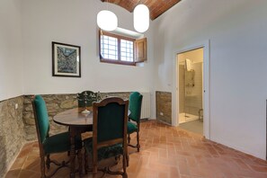 A dining area with a round table, two chairs, a framed painting, a window, and a shower room. - Le Mortinete Farmstay: Moraiolo (Toscana)