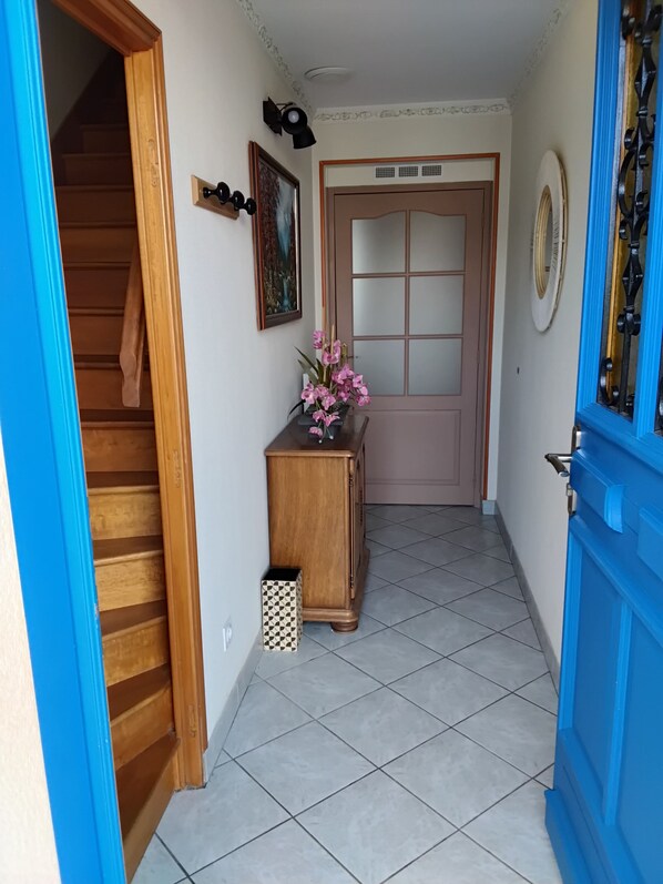 A hallway with a wooden staircase, a wooden cabinet with a vase of flowers, a door with a frosted glass panel, and a blue door with a decorative metal frame.
