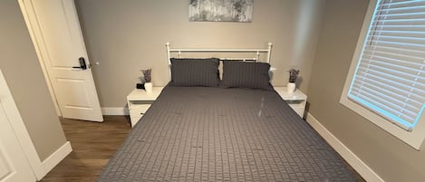 A bedroom with a bed, two bedside tables, a door, a window with blinds, and a wall-mounted artwork.