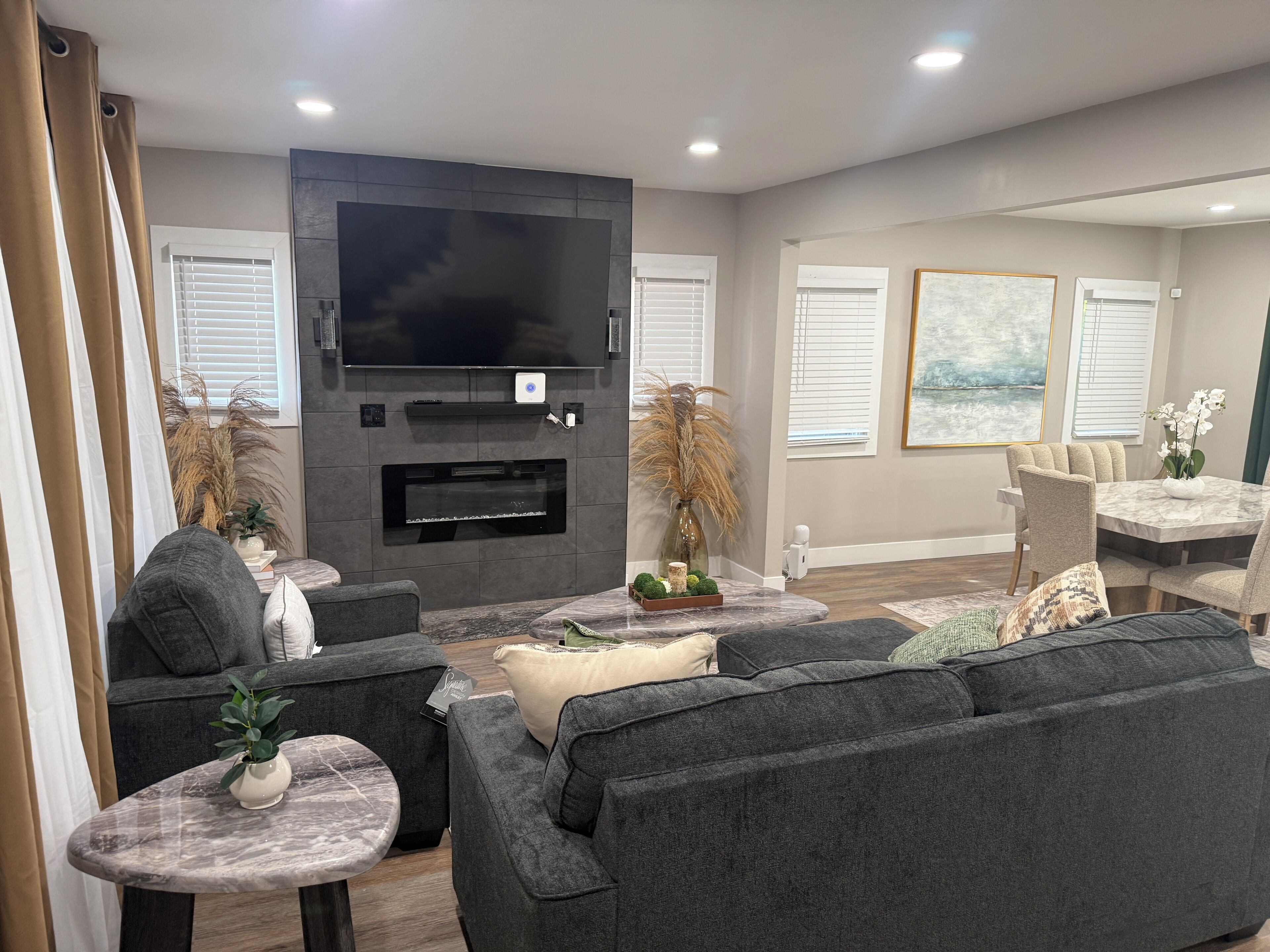 A modern living room with a flat-screen TV, fireplace, comfortable seating, and decorative plants.