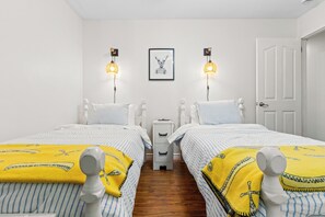 A twin bedroom with two beds, bedside tables, a nightstand, a framed picture on the wall, and a door.
