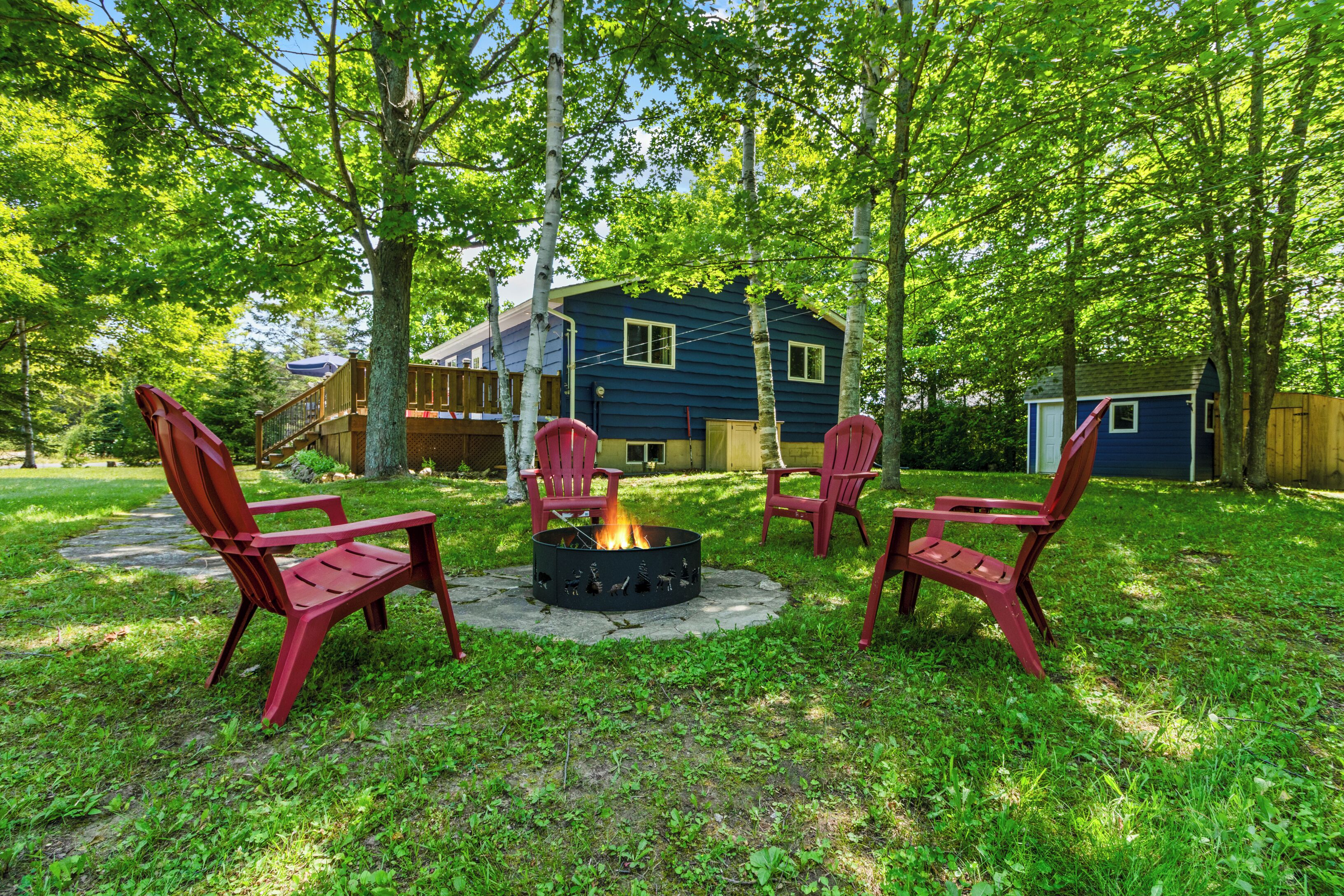 Un jardin avec un foyer entouré de chaises rouges, une maison bleue et une terrasse en bois.