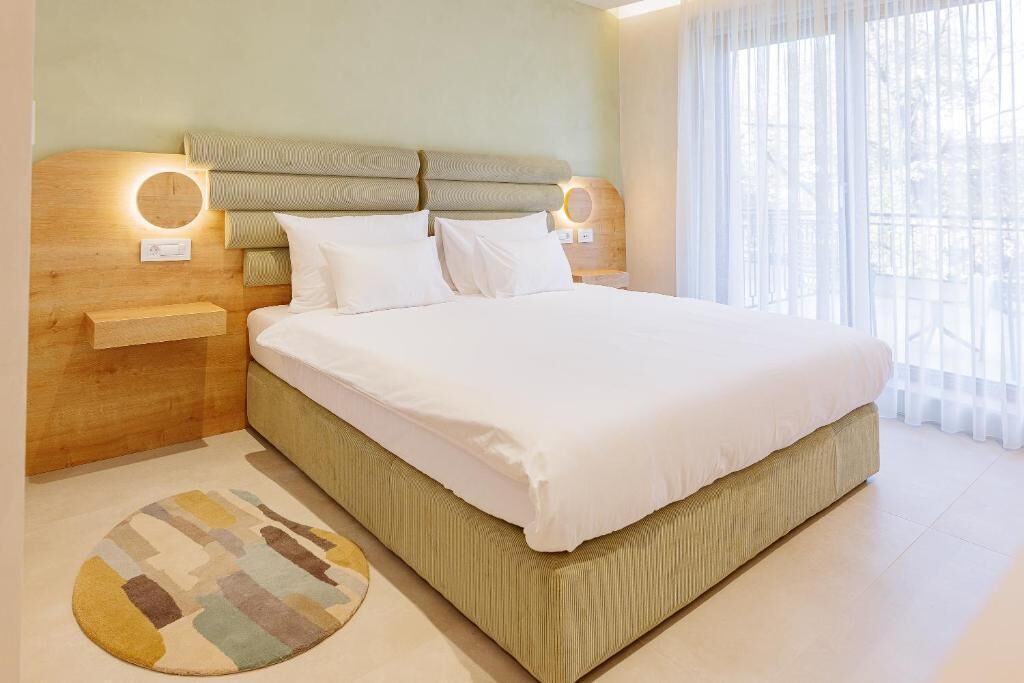 A modern bedroom with a large bed, a headboard with built-in lighting, a bedside table, and a view through a window.