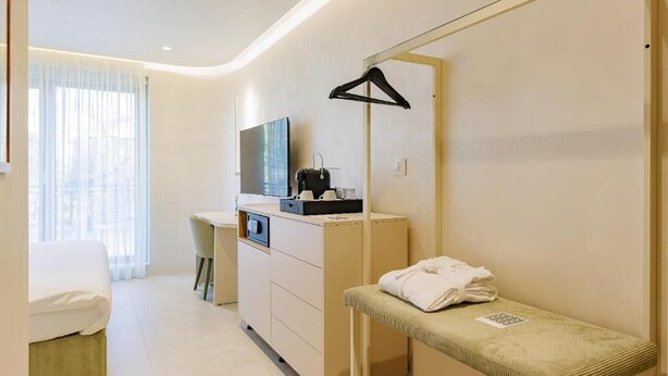 A hotel room with a bed, a desk with a chair, a TV, a mirror, and a bench with towels.