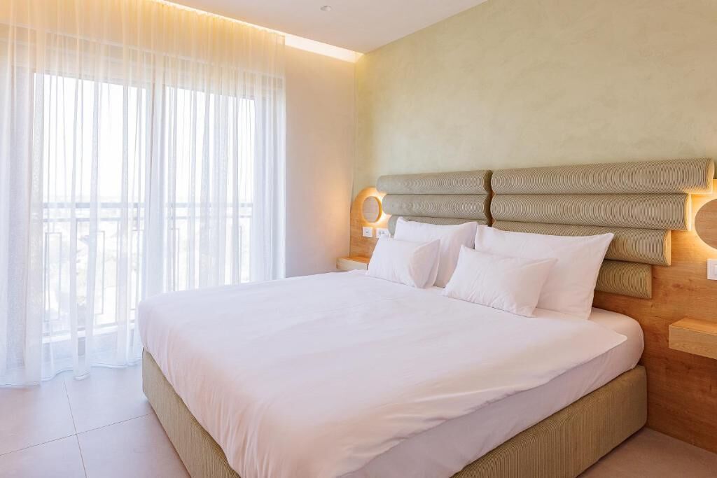 A hotel room with a large bed, a wooden headboard, and a view through sheer curtains.