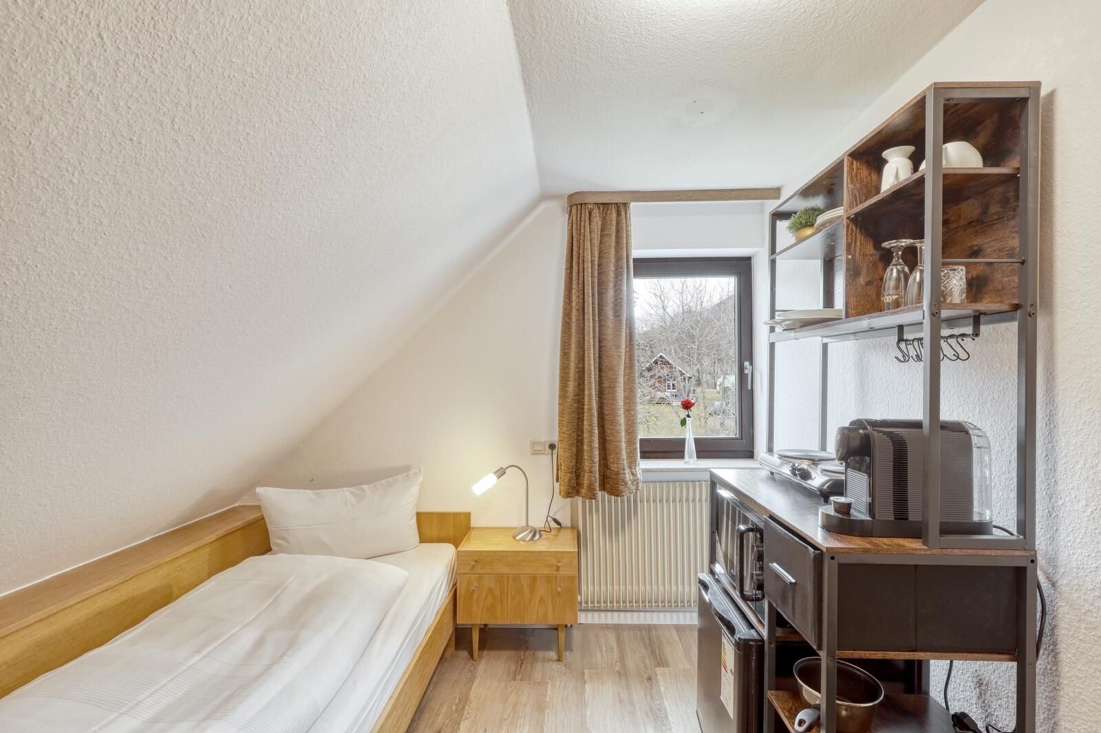 Una habitación pequeña y ordenada, con una cama individual, una mesita de noche, un escritorio con un hervidor y un radiador.