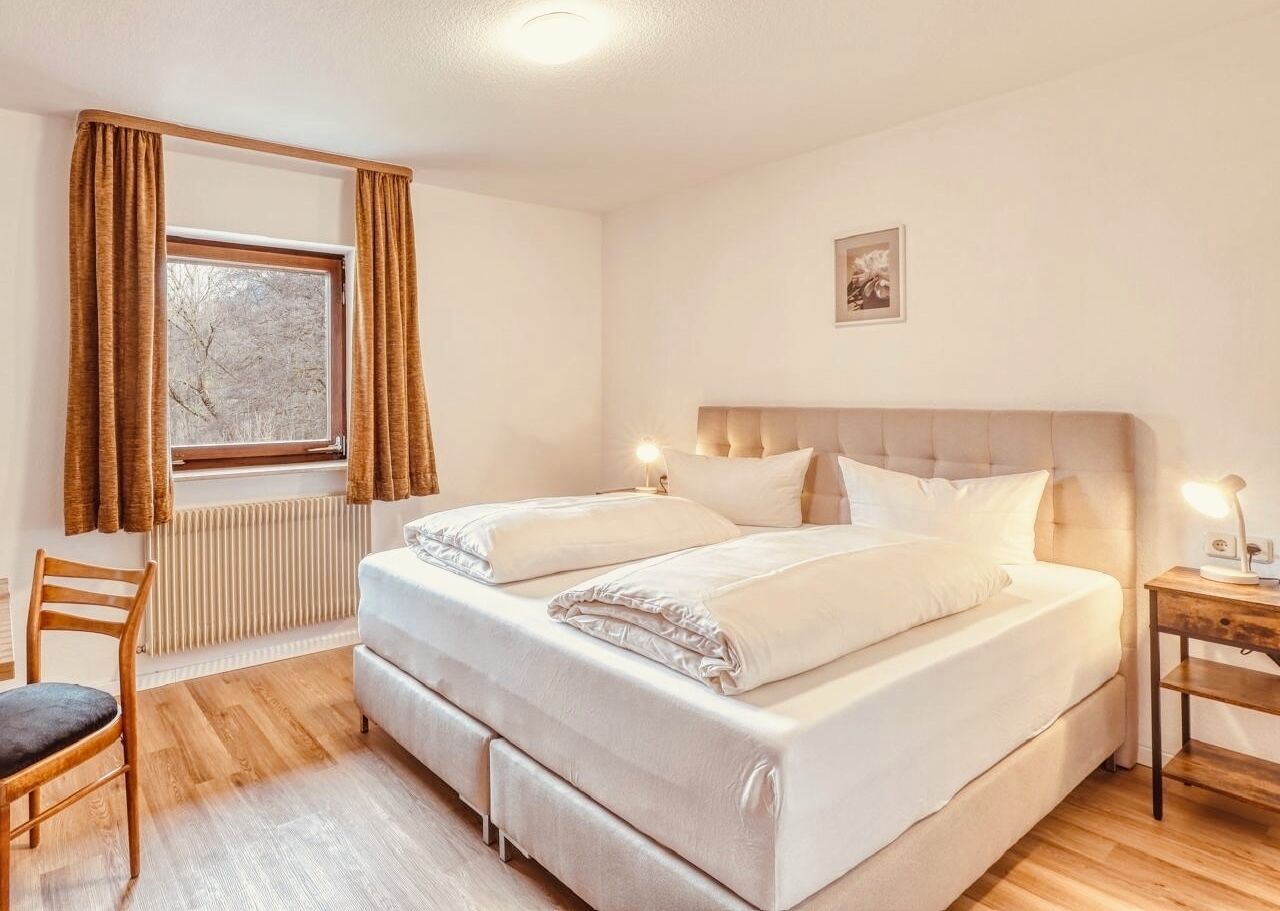 Une chambre à coucher avec un lit, une chaise, une table de chevet et une fenêtre avec des rideaux.