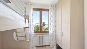Une petite pièce bien éclairée, avec un lit superposé, un radiateur, une fenêtre avec des rideaux et une armoire.