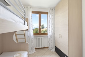 A small, well-lit room with a bunk bed, a radiator, a window with curtains, and a wardrobe. - Regarda - Basiliko Cosy Apartment with pool, pets allowed (Lazise)