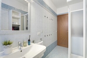 A bathroom with a white sink, a mirror, a shower, and a wooden door. - Regarda - Basiliko Cosy Apartment with pool, pets allowed (Lazise)
