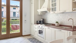 Une cuisine avec des meubles blancs, une cuisinière à gaz, un four et un évier. Il y a un balcon avec une chaise et une vue sur les bâtiments environnants.