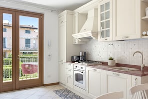 A kitchen with white cabinetry, a gas stove, an oven, and a sink. There is a balcony with a chair and a view of buildings outside. - Regarda - Basiliko Cosy Apartment with pool, pets allowed (Lazise)