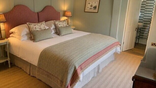 A neatly made bed with pillows and a blanket, a bedside table with a lamp, and a door leading to a staircase. - Super King/Twin Room at The Weld Arms (Wareham)
