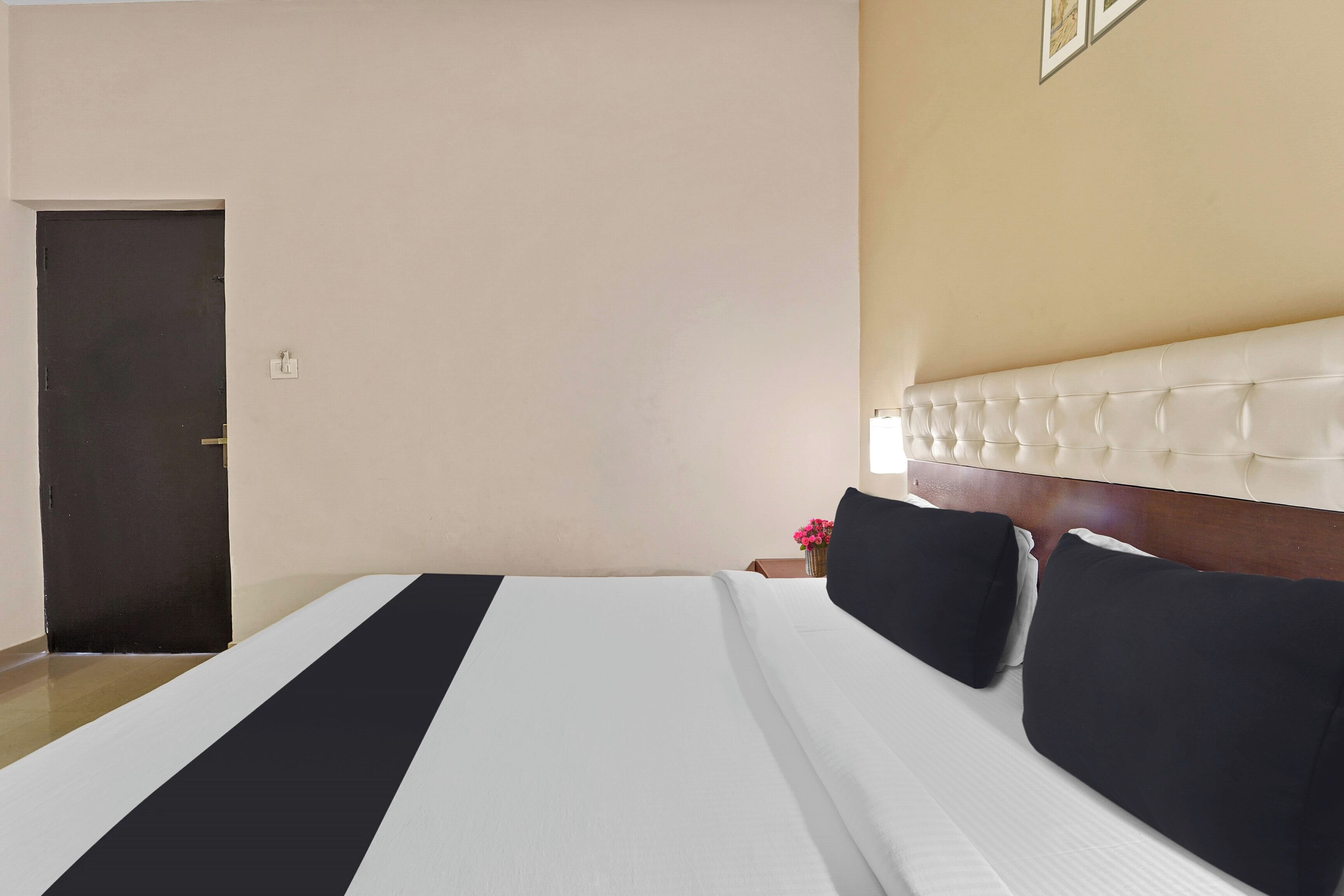 Una habitación de hotel con una cama, una puerta, una mesita de noche con un florero y una pared con un cuadro enmarcado.