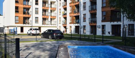 A modern apartment building with multiple balconies, a swimming pool, and a fenced parking area.