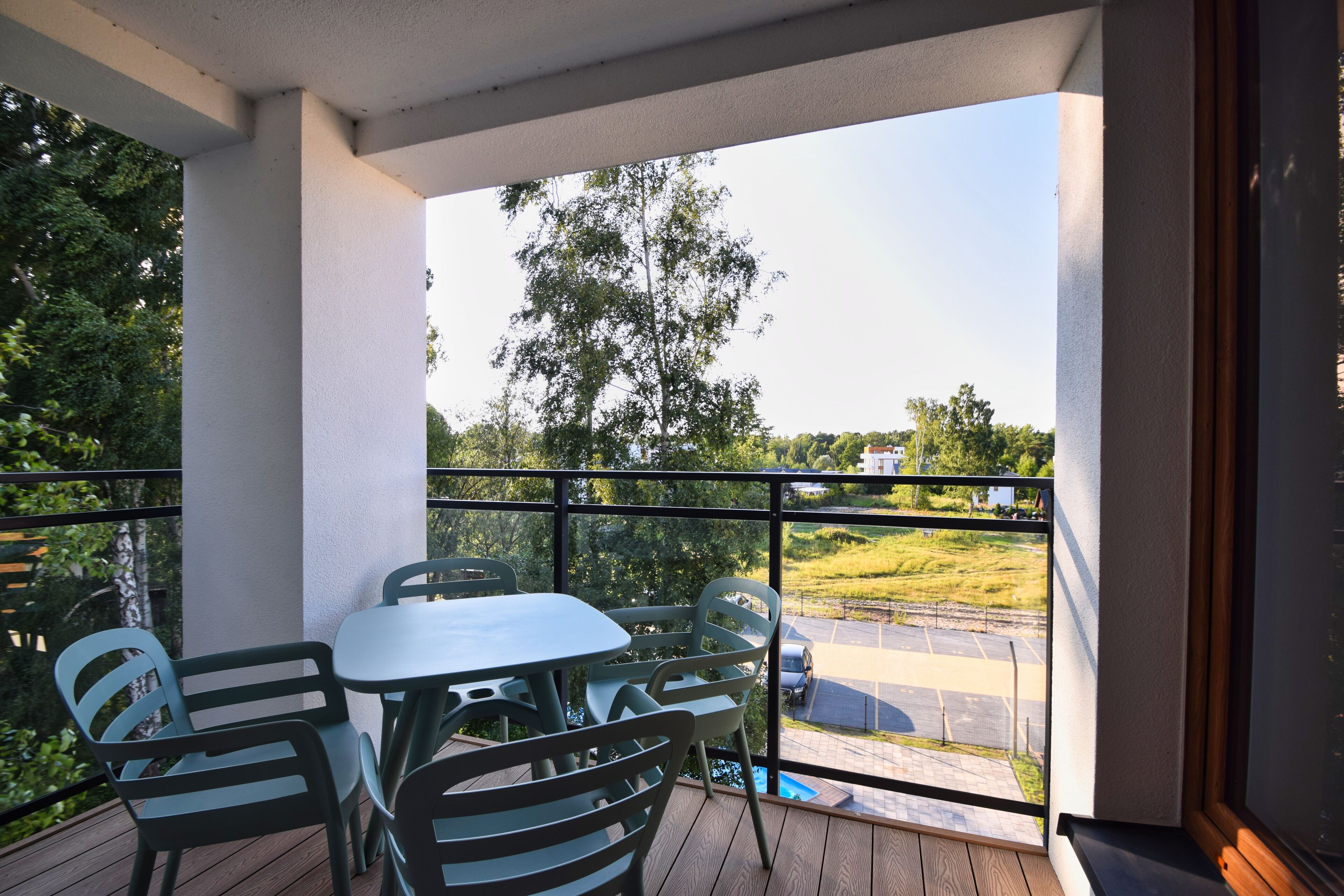 A balcony with a table and chairs, overlooking a parking area and greenery.