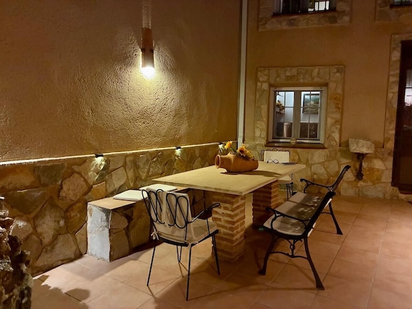 An outdoor patio with a stone wall, a table, two chairs, and a vase with flowers.