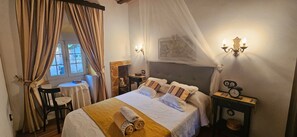 Un dormitorio con cama, mesitas de noche, una silla, ventana con cortinas y un cuadro colgado en la pared.