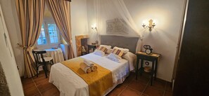 A bedroom with a bed, a bedside table, a chair, a window with curtains, and a wall-mounted light fixture. - Casa Turística Los Girasoles – Rural Getaway with Fireplace, Patios & Pool Access (Villarejo de Fuentes)
