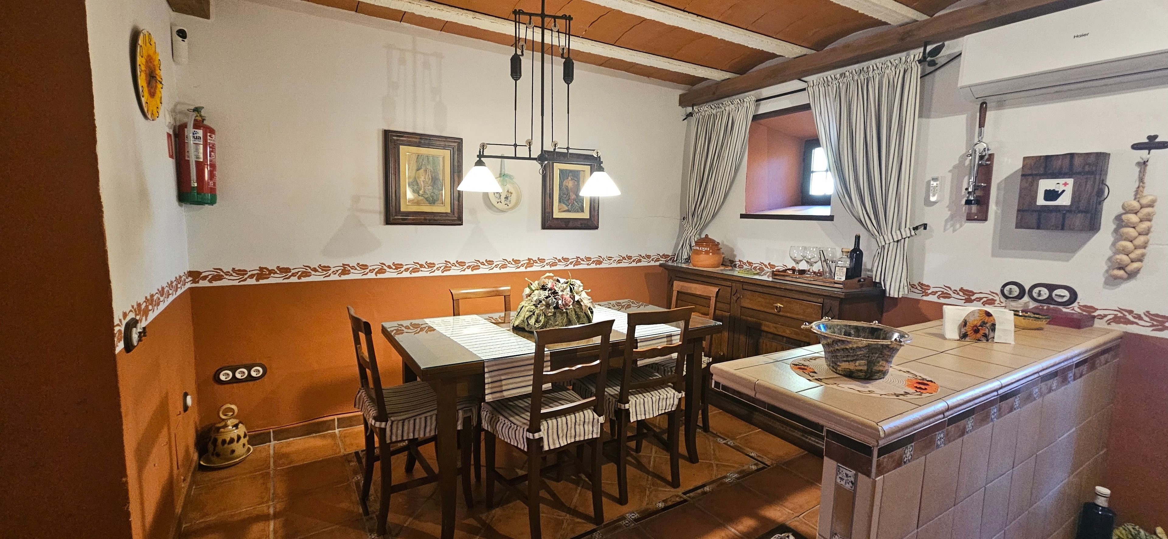 Un coin repas avec une table dressée pour quatre personnes, une cuisine avec une fenêtre et un luminaire suspendu.