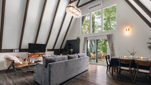 A modern living room with a grey sofa, wooden flooring, a television, and a dining area with a table set for two. - Luxury A-Frames - Pet-Friendly - Fenced Yard - Sleeps 12 - Minutes to Greenway! (Asheville)
