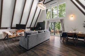 Ein modernes Wohnzimmer mit einem grauen Sofa, Holzboden, einem Fernseher und einem Essbereich mit gedecktem Tisch für zwei Personen.
