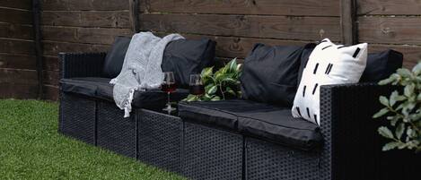 Un sofá negro para exteriores con cojines y una manta, junto con dos vasos de vino tinto y una planta en maceta.