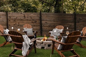 Ein Feuerkorb, umgeben von Adirondack-Stühlen mit Decken und Kerzen, vor einem Holzzaun.