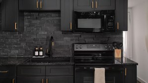 A kitchen with dark cabinetry, a black countertop, a microwave, a stove, and a brick backsplash. - Luxury A-Frames - Pet-Friendly - Fenced Yard - Sleeps 12 - Minutes to Greenway! (Asheville)