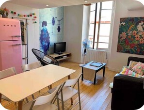 Un espace de vie compact comprenant un coin repas, une kitchenette, un téléviseur et une grande fenêtre laissant entrer la lumière naturelle.