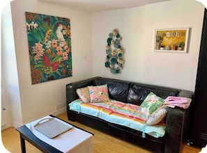A living room with a black leather sofa, colorful cushions, a white coffee table, a wall art piece, and a decorative wall hanging. - Port/Old Town: Quiet Duplex Loft (Nice)