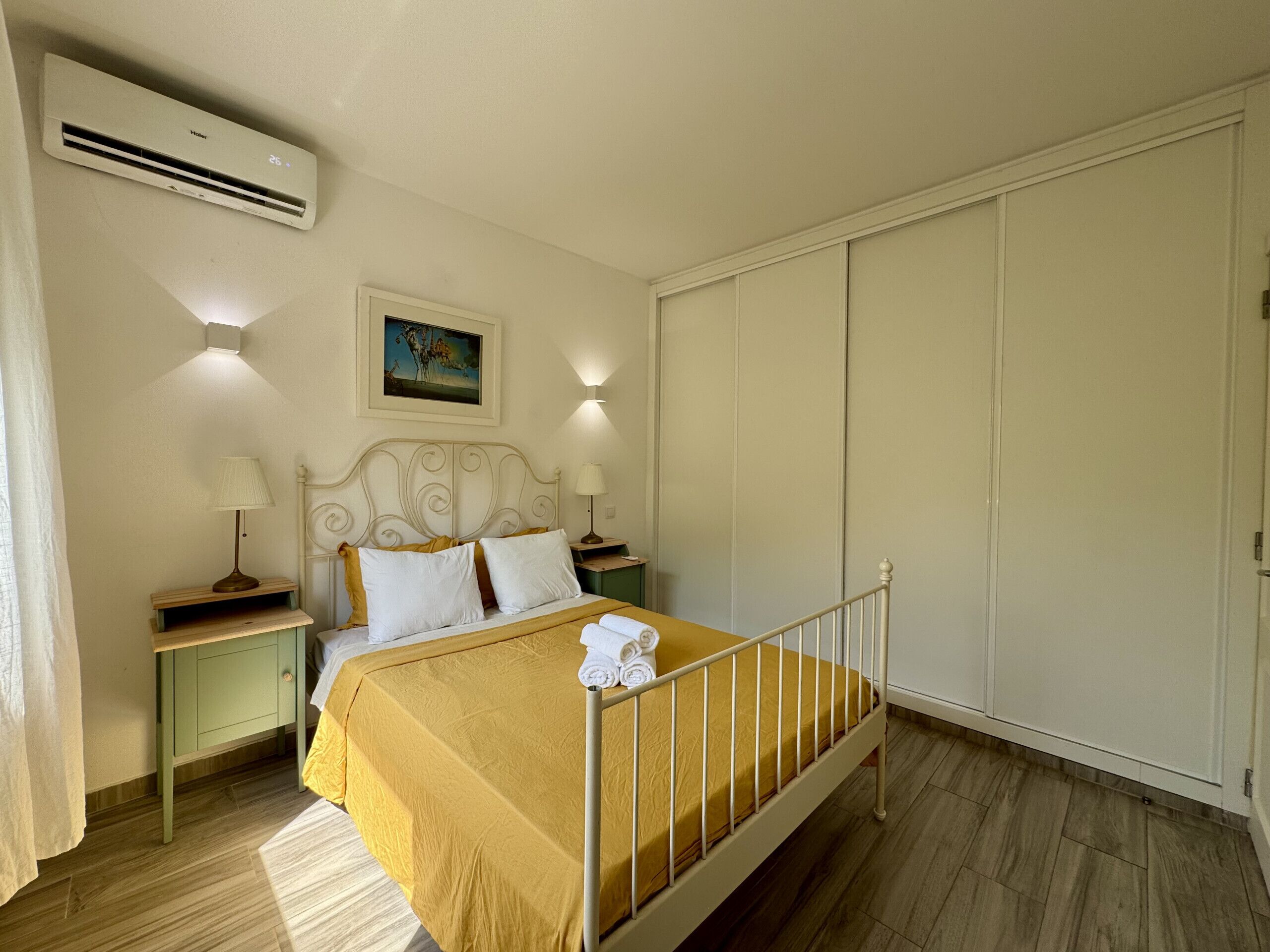 A bedroom with a bed, bedside tables, a lamp, a painting on the wall, and an air conditioning unit.