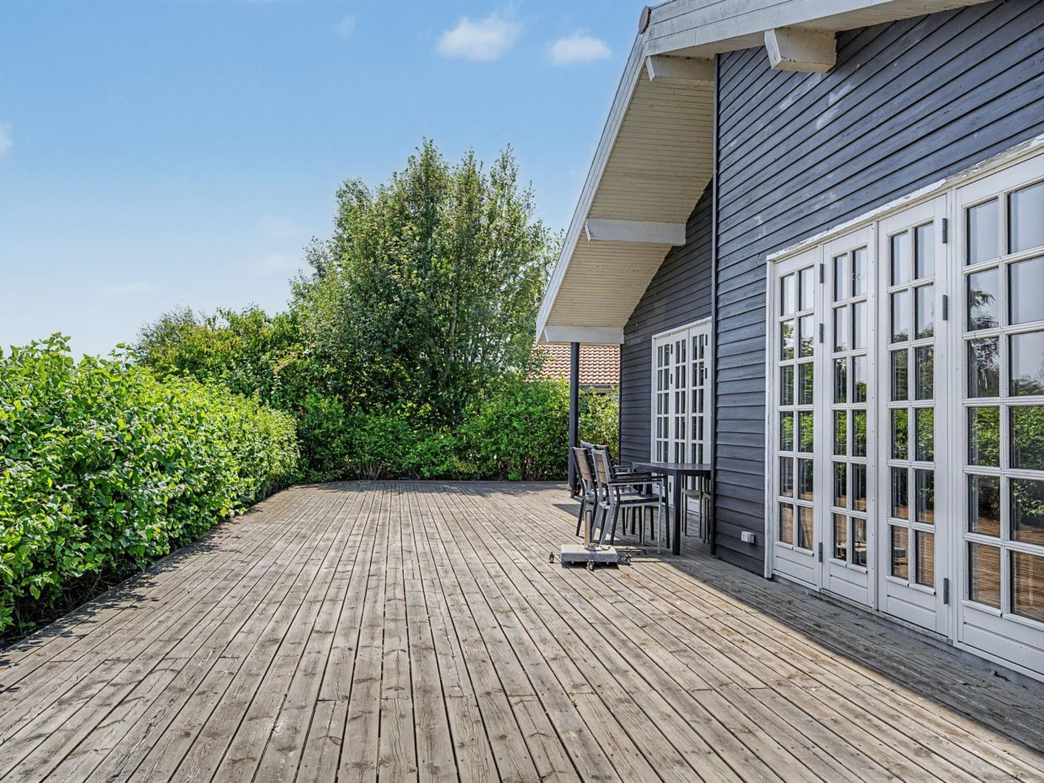 A wooden deck with a table and chairs, surrounded by greenery and a building with large windows.