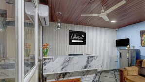 Ein Empfangsschalter mit Marmorplatte und Holzapplikationen, ein Deckenventilator, ein an der Wand befestigtes Fernsehgerät und ein Schild mit Firmenhinweisen.