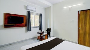 Ein Hotelzimmer mit Bett, Wandfernseher, Fenster mit Vorhängen, kleinem Tisch mit Wasserkocher und Tassen sowie einer Holztür.