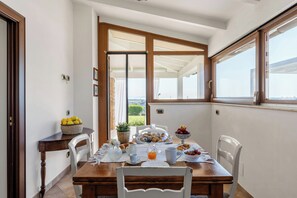 Zona pranzo con tavolo in legno, sedie bianche e vista sull'esterno attraverso una porta in vetro.