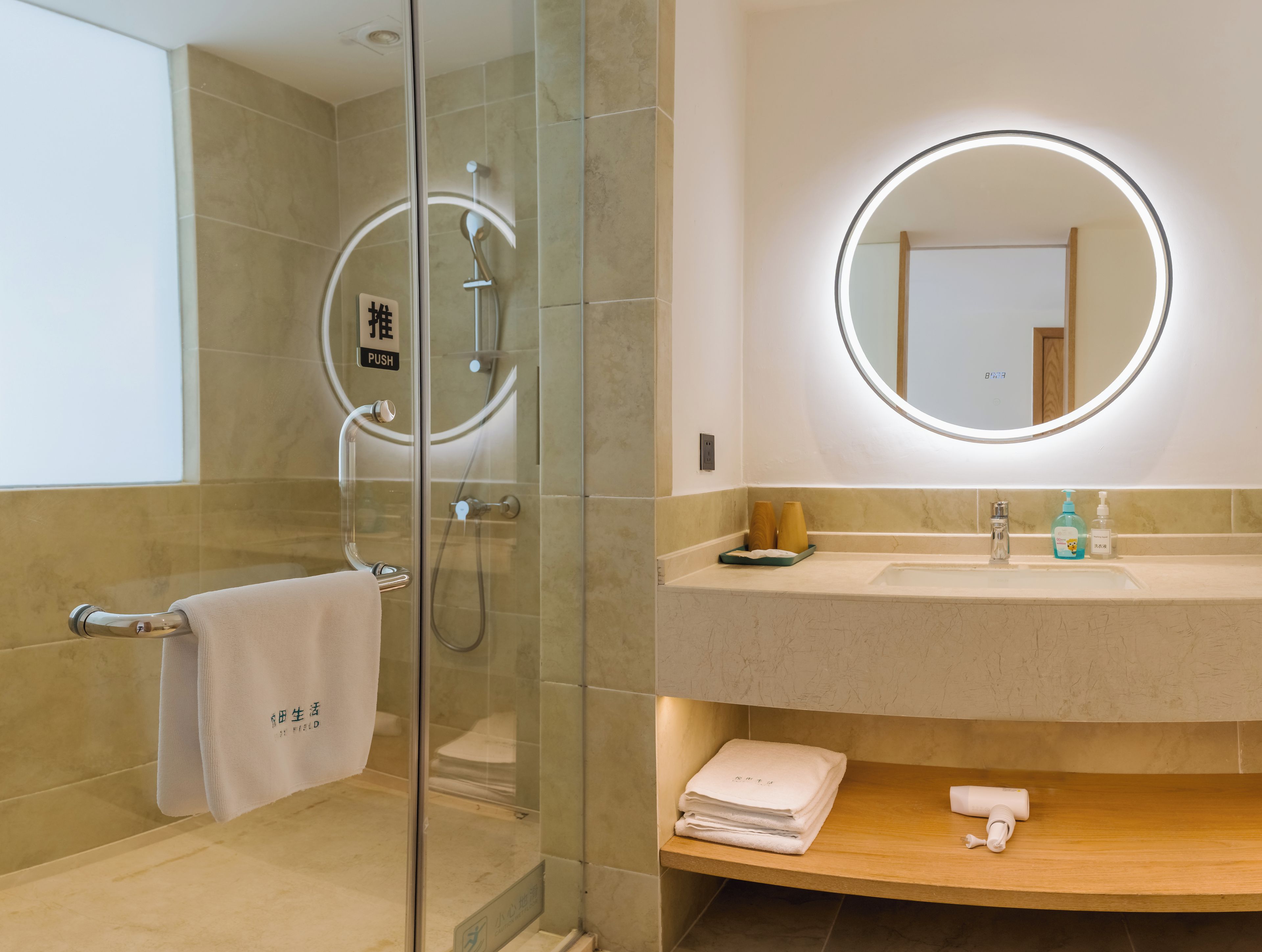 A modern bathroom with a glass-enclosed shower, a round mirror with a light, a sink with toiletries, and a towel rack.