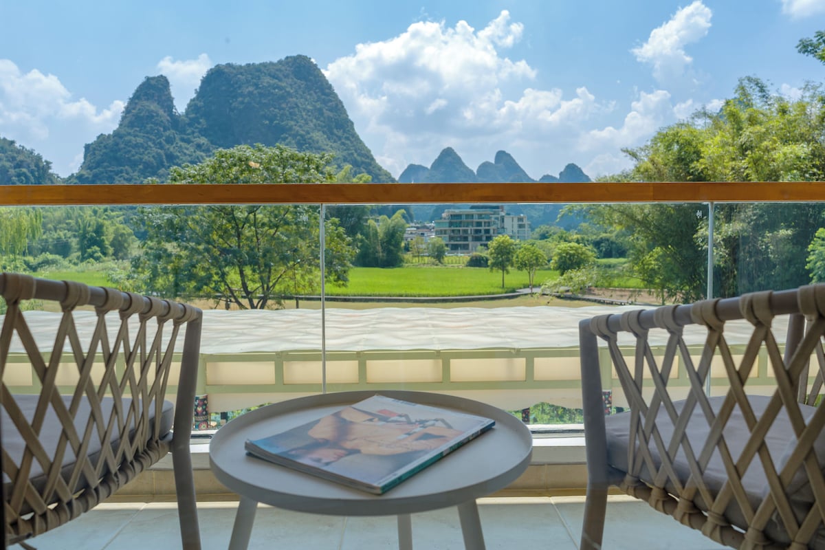 A balcony with wicker chairs, a small round table with a magazine, and a view of a mountainous landscape with buildings in the distance.
