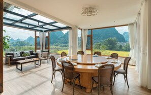 A dining area with a round table and chairs, large windows with curtains, a sofa, and a view of mountains. - Yangshuo YueTian Riverside Retreat - Boutique Stay by Yulong River - Talor Made Tours & E-bike Rental (Guilin)