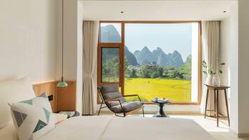 Une chambre avec un grand lit, une chaise, une petite table et une vue sur un paysage comprenant des montagnes et un champ.