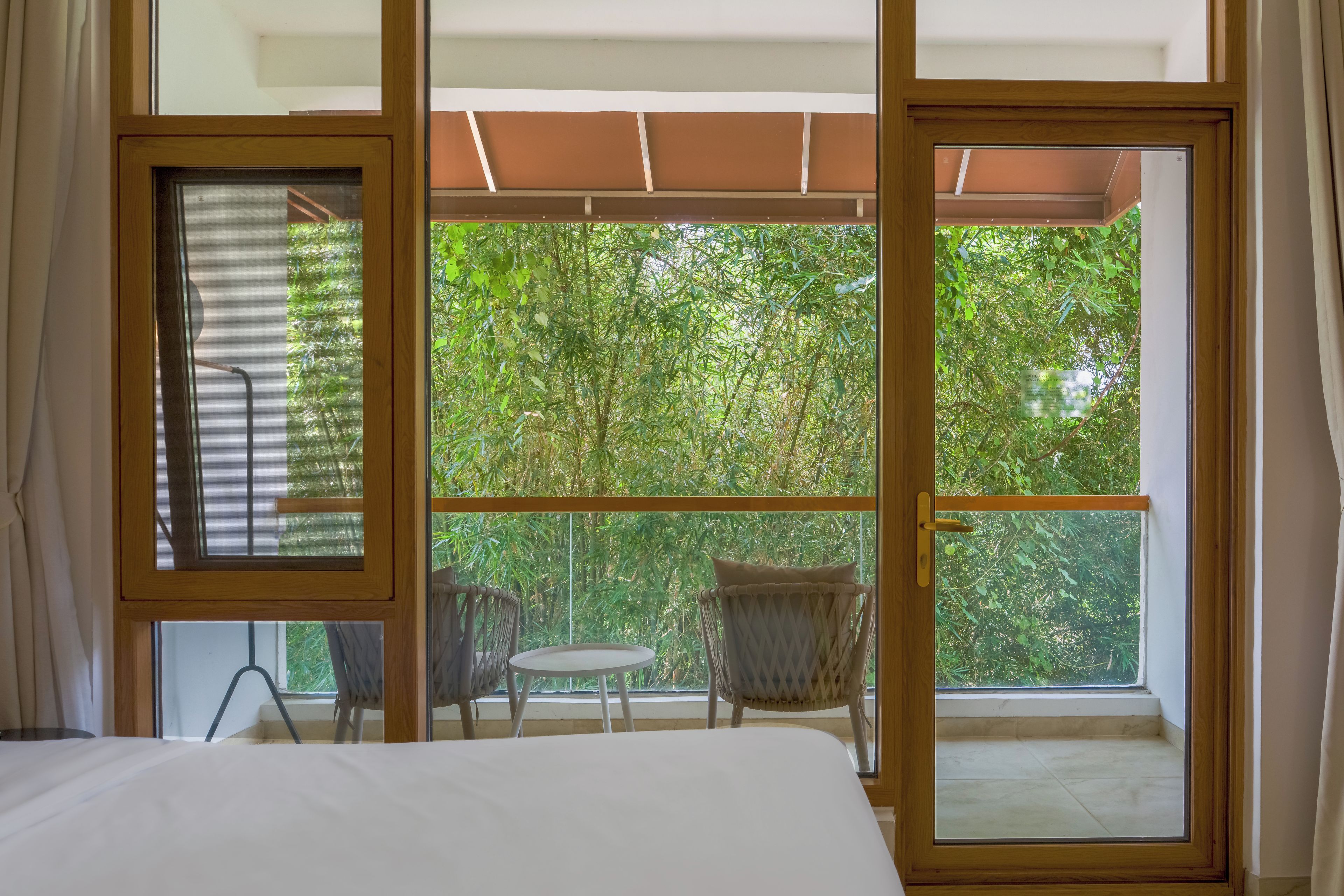 A room with a bed, a chair, and a table, with a view of greenery outside through large windows.